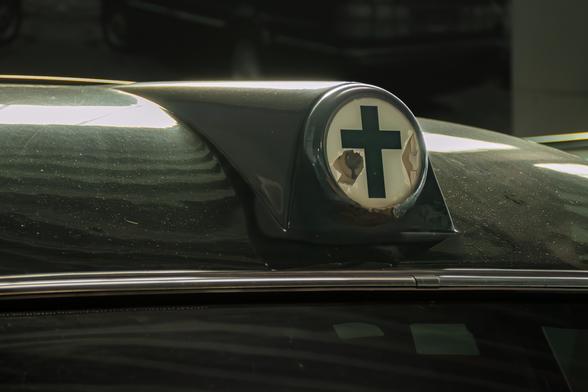 Dieses Bild zeigt eine Nahaufnahme des Daches eines dunklen Fahrzeugs, eines Leichenwagen-Oldtimers. Im Fokus steht eine runde, beleuchtete Anzeige oder ein Emblem, das ein dunkles Kreuz auf weißem Grund zeigt. Dieses Symbol ist leicht beschädigt, mit sichtbaren Rissen im Glas oder der Abdeckung, was dem Objekt eine gealterte oder beanspruchte Ästhetik verleiht. Die Oberfläche des Autos ist leicht staubig und reflektiert das Licht, was auf eine dunkle Umgebung oder eine längere Standzeit hindeutet. Die Szene wirkt melancholisch und konzentriert sich auf dieses spezifische, möglicherweise historische oder symbolische Detail am Fahrzeug.