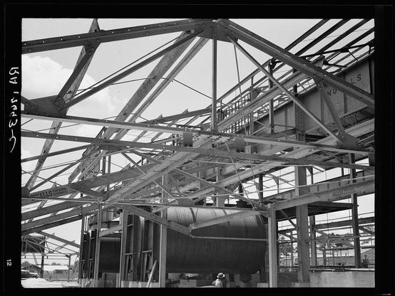 The image depicts a black and white photograph of an industrial construction site. It shows the skeletal framework of what appears to be a large ship or vessel, supported by numerous steel beams and girders. The structure is incomplete but substantial in scale, suggesting that it will house heavy machinery or serve as a massive storage facility.

In one corner of the frame, we can see part of a person wearing a hat, indicating human presence at the site. This individual seems to be standing on a platform or walkway designed for workers amidst such large-scale construction projects. The environment is open-air with daylight illuminating the scene, and there are no discernible details about the geographical location.

The image carries handwritten Japanese characters in the top left corner, which could indicate a date or some other form of documentation related to the photograph's context. However, without understanding the language or having additional information on this specific site, it is difficult to ascertain further specifics beyond what has been described above.