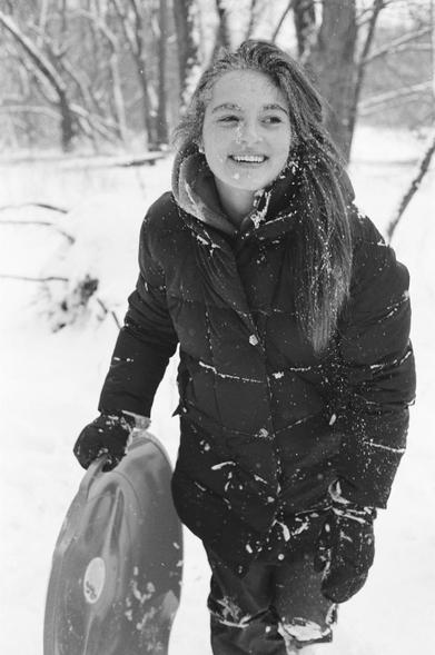 Black and white photo of a young woman smiling and holding a round sled. Snow dots her winter jacket, gloves, hair, and face—evidence of a sliding wipeout