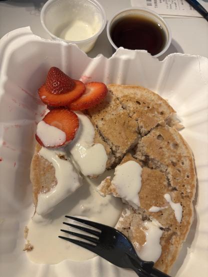 Takeout container, fork, pancake, ramekins of syrup and white sauce, sliced strawberries.