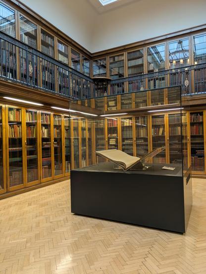 A large, open book is displayed in a glass case surrounded by bookshelves filled with numerous colorful books in a two-story library.