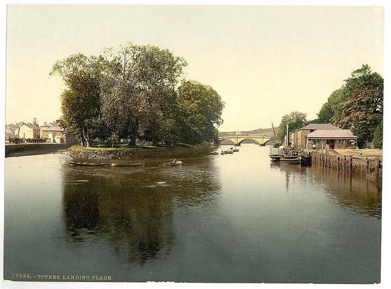 The image is a vintage postcard depicting the River Dart in Totnes, England. It features calm waters with reflections of surrounding trees and structures. In the foreground, there are boats docked at what appears to be a landing place or wharf. The middle ground shows another boat on the river, moving away from the viewer's perspective. To the left, dense greenery lines part of the riverside landscape. Across the river in the background is an arched bridge connecting two parts of Totnes across the Dart River.
A wooden structure with a thatched roof and several windows can be seen to the right side on land near the water’s edge. Various buildings are visible along both sides of the river, indicating it's likely a busy area for commerce or transportation during the time this photo was taken. The sky is overcast, suggesting an early morning or late afternoon setting with subdued lighting conditions.
The overall atmosphere conveyed by the image evokes historical charm and simplicity from the turn-of-the-century period in England.