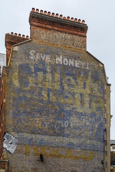 Tall gable-end wall with chimney stacks on top. The bricks are covered in a medley of overlapping fading painted signs, with "Save Money, Gillette" being most prominent.