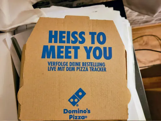 A brown cardboard Domino's pizza box sits atop a stack of papers. Blue text on the box reads, "HEISS TO MEET YOU," followed by "VERFOLGE DEINE BESTELLUNG LIVE MIT DEM PIZZA TRACKER," and the Domino's Pizza logo at the bottom.