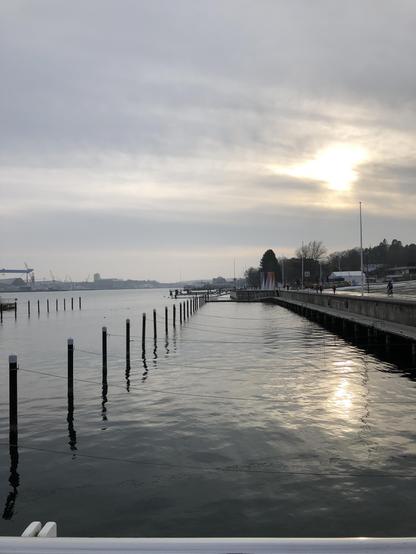 Verzerrte Sonne in dunstigen Wolken über der dem ruhigen Fördewasser am leeren Boots-Hafen.