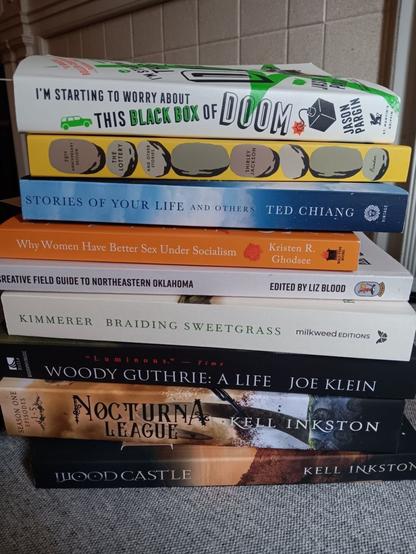 A pile of paperbacks. From top to bottom: I'm Starting To Worry About This Big Black Box of Doom by Jason Pargin, The Lottery and Other Stories by Shirley Jackson, Stories of Your Life and Others by Ted Chiang, Why Women Have Better Sex Under Socialism by Kristen R. Ghodsee, Creative Field Guide to Northeastern Oklahoma ed. by Liz Blood, Braiding Sweetgrass by Robin Wall Kimmerer, Woody Guthrie: A Life by Joe Klein, and Nocturna and Woodcastle by Kell Inkston