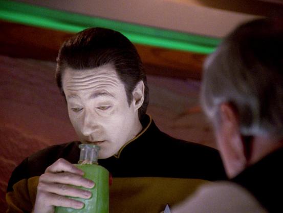 A scene from Star Trek: The Next Generation shows the character Data holding a green bottle close to his mouth while seated at a table. He is wearing a black and gold Starfleet uniform. Another person with gray hair is visible in the foreground, facing Data, but their face is partially out of focus. The background features a softly lit wall with a green light strip above it.