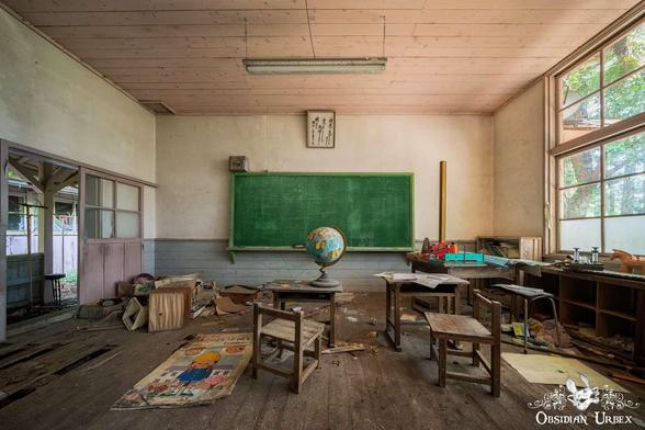 A decaying classroom with scattered wooden furniture, a large green chalkboard, and a globe. On the floor, a children's illustration lies near other debris.