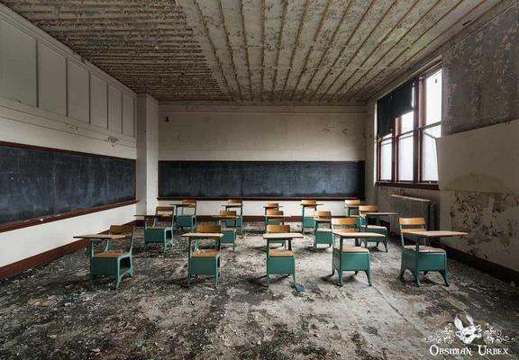An abandoned classroom features peeling paint, rows of teal desks with wooden tops, and two long chalkboards. The floor is covered in debris, hinting at disuse