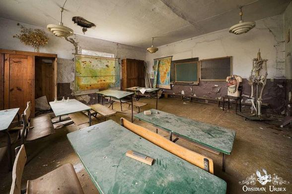 An abandoned classroom features worn green-topped desks, maps, blackboards, and skeletal models amidst peeling paint and a damaged ceiling.