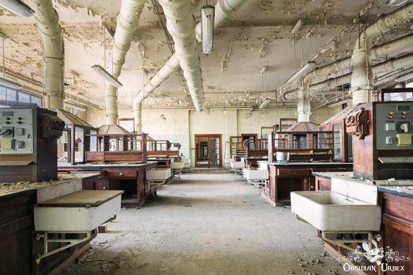 An abandoned, decaying laboratory classroom features peeling paint, old wooden benches with sinks, and exposed pipes