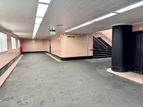 Corridor with pink tiled walls, a rounded black column to one side and stairs in the middle background going upwards. Overhead fluro lighting.