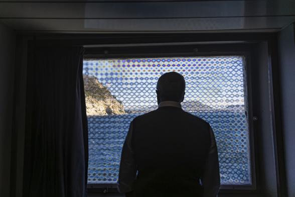 A passenger on a ferry boat is looking at the maritime landscape of the Bay of Naples through a window covered with a transparent shield decorated with geometrical patterns.