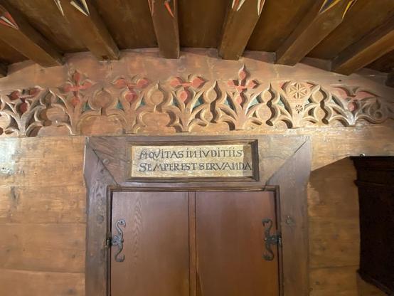 Eine Holzwand mit kunstvollen Schnitzereien und farbenfrohen Designs über einer geschlossenen Tür. Darüber ein hölzernes Tonnengewölbe. Über der Tür ist in den Rahmen eine lateinischen Inschrift eingelassen: "ÄQVITAS IN IVIDITIIS SEMPER EST SERVANDA".