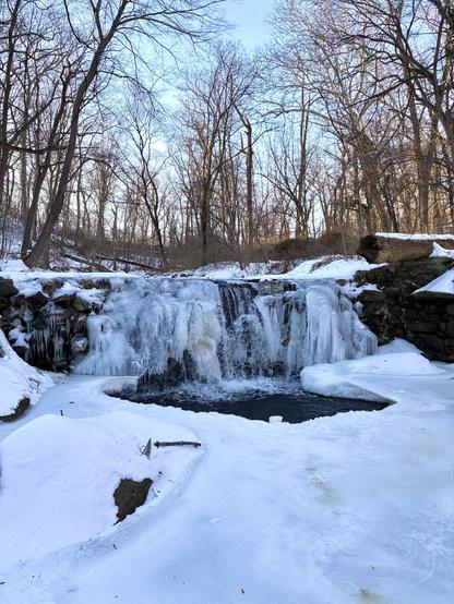 The waterfall is frozen except for the middle, where plunging water is still visible. The pool below is also frozen, with only the center open where the water crashes down. It’s a partly sunny late afternoon in the woods.