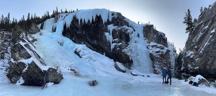 Photo of a rock face with a frozen waterfall cascading down in various places. One of the frozen waterfalls has an ice-climber part-way up, with a person standing on top of the waterfall, and two people standing below.