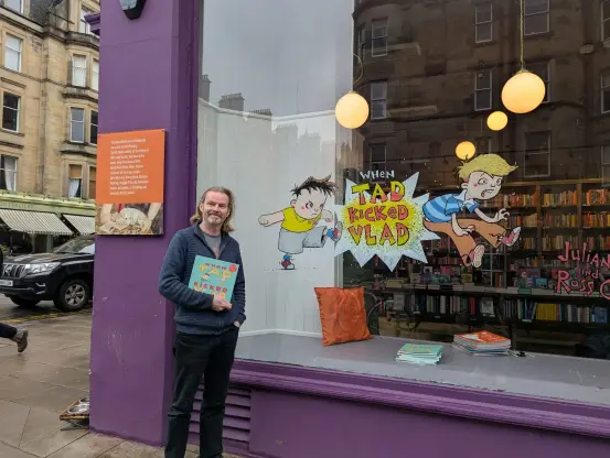 Ross Collins standing with his new picture book, outside the Edinburgh Bookshop with his recently completed window painting featuring characters from the book