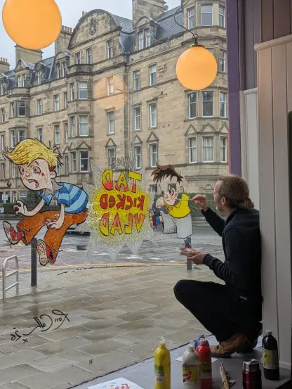 Illustrator Ross Collins kneeling in the bookshop window, painting in the colours of the outlines he already drew of his picture book characters