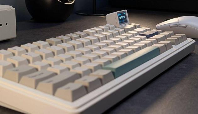 Close-up of a sleek, beige mechanical keyboard with a small digital display, next to a white computer mouse on a dark surface.