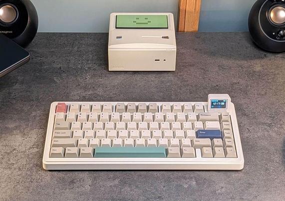 A vintage-style mechanical keyboard with cream-colored keys and a small display, placed on a textured surface in front of a retro mini PC.