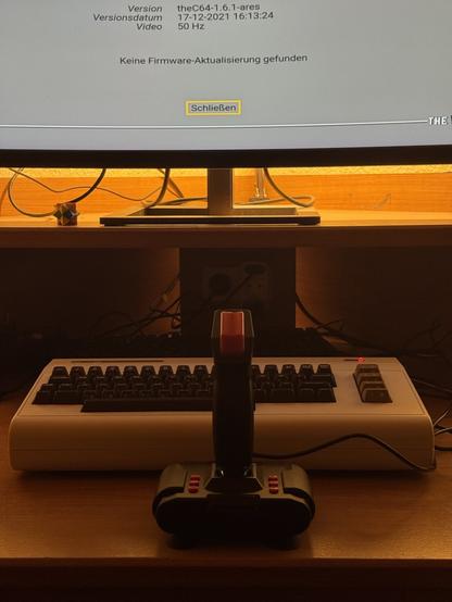An image of a retro computer setup featuring a Commodore 64 keyboard, a joystick controller, and a monitor displaying system information in German. The background has soft lighting, and there is a small geometric object on the desk.