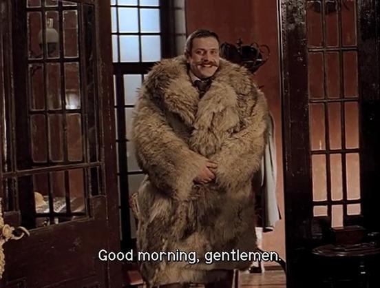 A grinning, richly mustachioed man stands in a doorway, dressed in a fur coat the size of a wardrobe, subtitled “Good morning, gentlemen”.