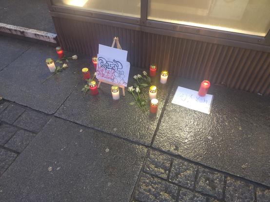 An einem Parkhauseingng im Regen stehen weiße und rote Grablichter. Es liegen einige Blumen dabei und Freunde von Manuel haben Schilder gemalt.