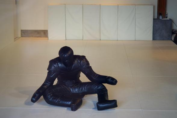 A life sized, black leather grappling dummy tiredly rests on the floor after practice, looking exhausted and forlorn in the quiet room.