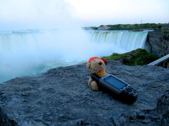 Das Bild zeigt einen Teil der Niagarafälle, aufgenommen aus einer Perspektive, die die Majestät der Wasserfälle hervorhebt. Im Vordergrund befindet sich ein felsiger Vorsprung aus grauem Stein, der mit kleinen Grashalmen und Moos bewachsen ist. Auf diesem Felsen sitzen zwei Objekte: ein kleiner, hellbrauner Teddybär mit einem orangefarbenen Hut und einem gelben Halstuch, und ein dunkler GPS-Gerät, das neben dem Bären liegt. Der Teddybär ist nach rechts geneigt, während das GPS-Gerät diagonal zum Teddybären positioniert ist.
Der Hintergrund wird von den beeindruckenden Niagarafällen dominiert, die sich über die gesamte Breite des Bildes erstrecken. Das Wasser stürzt in breiten Kaskaden von einer Felskante herab und bildet eine dichte Wolke aus Gischt, die einen großen Teil des mittleren und linken Bereichs des Bildes einnimmt. Die Gischt verleiht der Szene eine leicht dunstige, ätherische Atmosphäre. Das Wasser unterhalb der Fälle hat eine türkisfarbene bis blaugrüne Tönung und ist durch die Wellenbewegungen und die Gischt unruhig.
Am oberen Horizont sind auf der anderen Seite der Fälle Bäume und Gebäude zu erkennen. Die Bäume bilden einen dichten grünen Streifen, während einige Gebäude, darunter ein größerer, heller Bau und ein schmaler Turm, durch die Dunstschicht hindurch sichtbar sind. Der Himmel ist ein sanfter Übergang von blassem Blau zu Grau, was auf eine Dämmerung oder einen bewölkten Tag hindeutet.