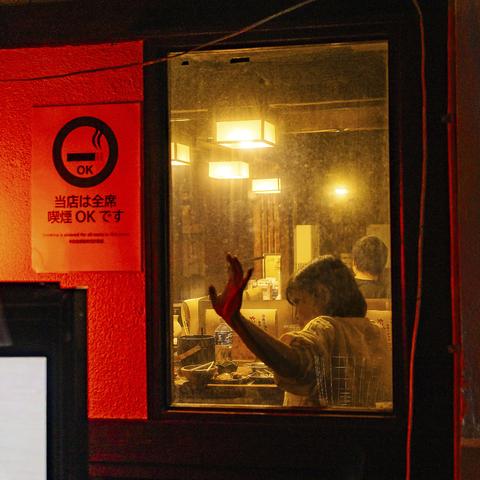 A dark street with a red lighting on a restaurant sign reading 'smoking allowed', inside the restaurant behind the window close to the sign, a lady his brandishing her lit cigarette to the window while talking to someone that can't be seen opposite her. The restaurant interior is lit in a warm color.
