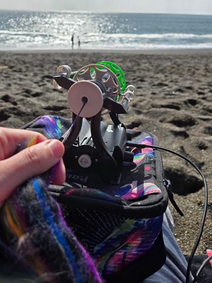 A hand spinning colorful roving on an EEW Nano perched on a lunch box, at the beach by the edge of the water.