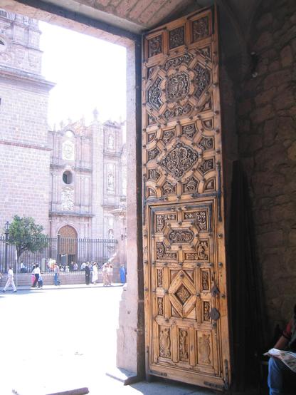 Die Fotografin steht in der dunklen Eingangshalle des Rathauses von Morelia in Mexiko und schaut durch das geöffnete große Tor auf die Straße und die Kirche gegenüber. Alles, was draußen ist liegt in grellem Sonnenlicht. Bei dem schweren Tor handelt es sich um eine koloniale Kassetten-Arbeit nach spanischem Stil. Im Tor ist eine kleinere Türe eingefügt, das man früher für Menschen öffnete. Das ganze Tor ist wahrscheinlich fünf Meter hoch und wurde für Pferdekutschen geöffnet, als es noch keine Autos gab.