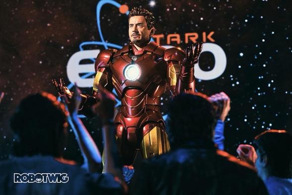 Tony Stark stands on a stage in front of a crowd of people in his iron man suit without his helmet. Stark Expo logo in background.