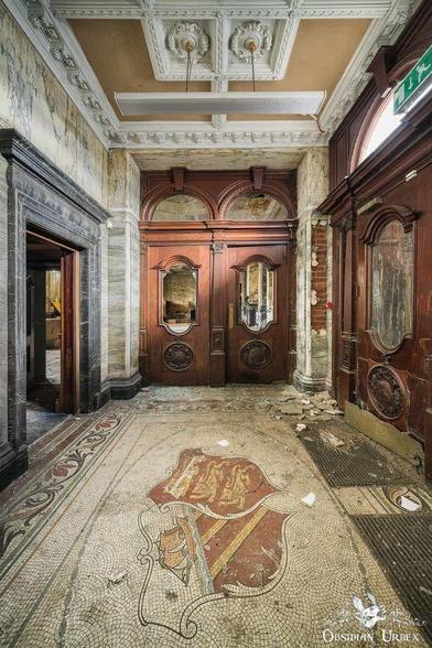 An ornate, abandoned bank hallway features a mosaic floor with a crest, dark wooden doors, marble walls,
