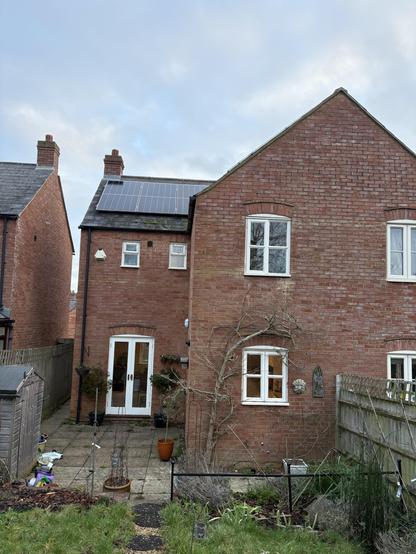 A two-story red brick house with a sloped roof. Solar panels are mounted on the roof. The backyard features a stone patio, several potted plants, and a garden area along with a wooden shed. There are two sets of windows with white wooden frames.