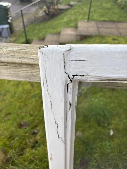 Close-up of a damaged white window frame with visible cracks and peeling paint. In the background, there is greenery and a pathway.