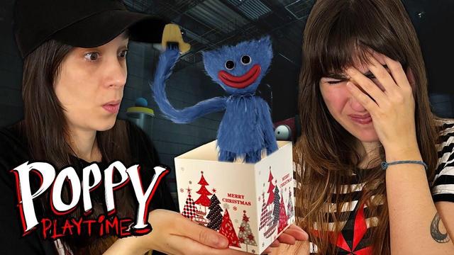 Dunkel sorprendida con un regalo de navidad del que sale Huggy Wuggy. Rena tiene los ojos cerrados y se lleva la mano contra la cara.