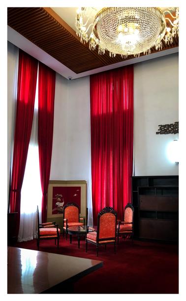 A vertically oriented photo of a room with elegant decor, with red the dominant color. Rich red wall-to-wall carpeting covers the floor. Four chairs with carved wooden frames, arched backrests, and red upholstery are arranged around a low round table with a reflective top. Behind the chairs is a two-panel folding screen with an image of white cranes against a red sky. Two tall windows with red curtains soar up to meet the high ceiling: the right with its curtains drawn, the left with its curtains tied off to the sides to let the sunlight in. An elaborate crystal chandelier hangs from the ceiling, surrounded by a broad border of wooden slats. At right is a wooden display case under a bright wall-attached lamp and a mounted wall decoration. A rectangular table in the foreground pushes in at lower left, its glossy surface reflecting the sunlight.