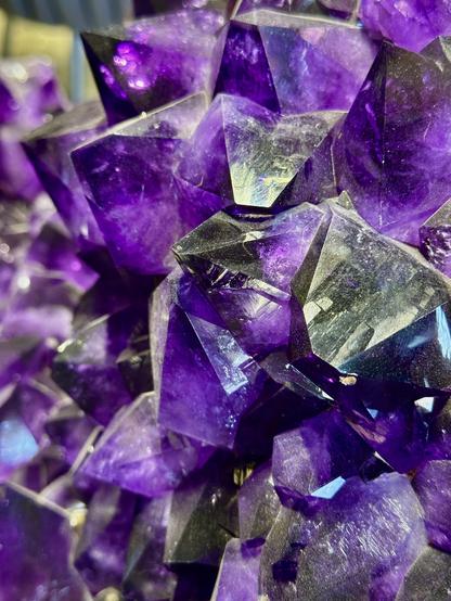 Closeup of large amethyst crystals in a large vug displayed on its back due to the massive size as big as a person lying down and holding their knees in close. This view shows large crystals that are light to dark purples. Crystals are about 1-2” at widest and about 1 to 1 1/2” tall from base to points. They are mostly the characteristic deep purple with deep bluish purple to warm reddish purple. If the specimen is exposed to too much sunlight most of the time it will fade.