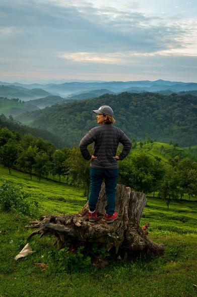 A person stands on a tree stump, facing away from the camera, with hands on hips. They are wearing a gray cap, a dark jacket, and hiking pants, overlooking a lush green landscape with rolling hills and misty mountains in the background.