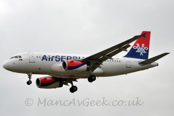 Side view of a twin engined jet airliner flying from right to left at a low altitude, with undercarriage lowered, and flaps extended from the front and rear of the wings, suggesting it is about to land.
The plane is largely white, with blue "Air Serbia" titles on the side of the forward fuselage, the last few letters partially obscured by the wings.
These titles are repeated, inverted, in red, under the wing-box.
The engine pods mounted under the wings are red on top, with a thick dark blue stripe running from the bottom at the front to the top at the rear, then white on the bottom.
The tail is red on the top half and blue on the bottom, with white lines creating an image of a 2-headed eagle with blue chest markings.
Flat grey cloud fills the rest of the frame.