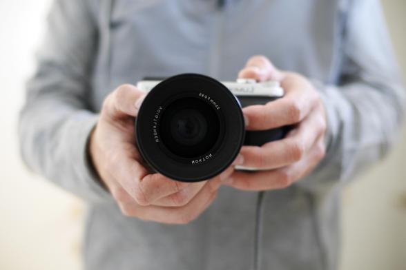 A picture of me holding my camera as seen in a mirror. The camera is in the center of the picture, facing directly forward towards the mirror. The camera is a Panasonic Lumix GX seven with a Voigtlander Nokton 25 mm F/0.95 lens (the text on the lens is backwards because of the mirror). Behind the camera you can see me in a gray shirt, blurred out of focus. The picture is framed such that you cannot see my head or my lower body.