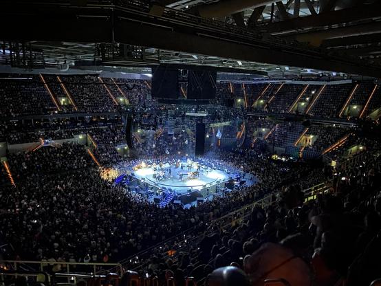 Ein Konzert in einer großen Arena mit einem kreisförmigen Bühne, umgeben von vielen Zuschauern, die das Event mit Handys dokumentieren.