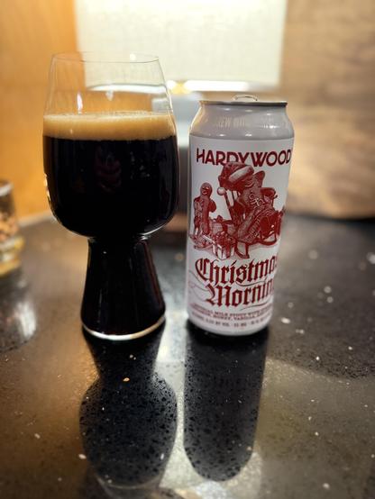 A stout glass full dark beer is next to a pint can with a white label picturing a gingerbread man in a rocking chair in shades of red