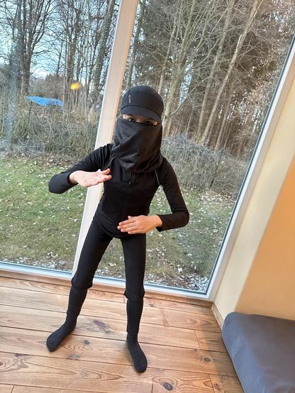 Kind in angedeuteter Verteidigungshaltung ganz in Schwarz mit Tuch um Mund und Nase sowie Käppi. Nur die Augen sehen aus schmalen Schlitzen hervor.