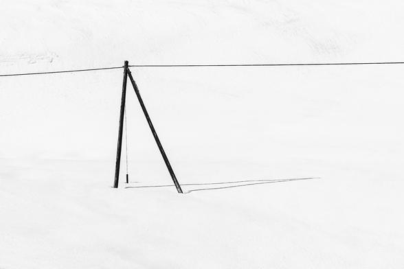 A minimalist black and white image featuring a single wooden support pole with a horizontal wire. The pole stands in a vast expanse of white snow, creating a strong contrast and emphasizing the simplicity of the scene.