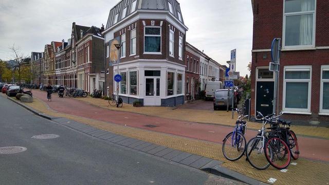 A red two-way cycle track passes the end of a little side street with three story Dutch homes on both sides. The access to the side street from the main road is via a steep ramp which crosses the cycle track and  buff coloured footway. There are soke bikes parked against a sign post to the right of the access.