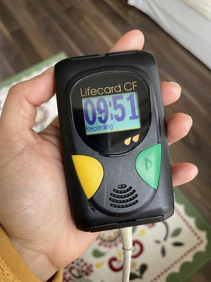 Holding the ECG recording box in my hand. It’s a chunky black plastic box with a simple LCD screen showing the time, and two big buttons. It looks like a 1990s pager or dumb phone.