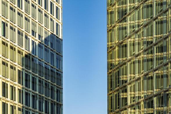 Image features two modern buildings with glass facades reflecting sunlight, creating an interplay of light and shadow, against a clear blue sky.