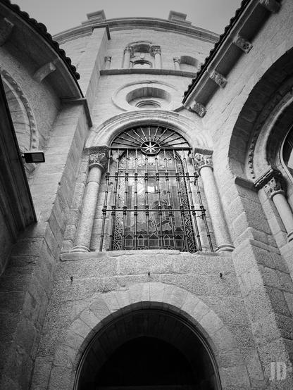 La imagen muestra la fachada de un edificio de piedra de estilo histórico, probablemente una iglesia o catedral. Se observa un arco de entrada en la parte inferior y, sobre él, una gran ventana con vidrieras decorativas y una reja de hierro forjado. La estructura presenta columnas adosadas y detalles ornamentales, con un aspecto macizo y simétrico. La perspectiva está tomada desde abajo, enfatizando la altura del edificio. La imagen está en blanco y negro, lo que resalta el contraste y la textura de la piedra.